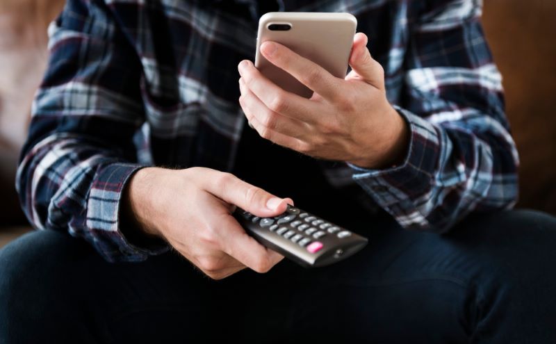 Uma pessoa sentada segura o controle remoto da TV numa mão e o celular na outra.