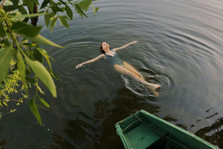 Uma mulher boia sobre a superfície da água escura, próxima a um barco e a uma folhagem.